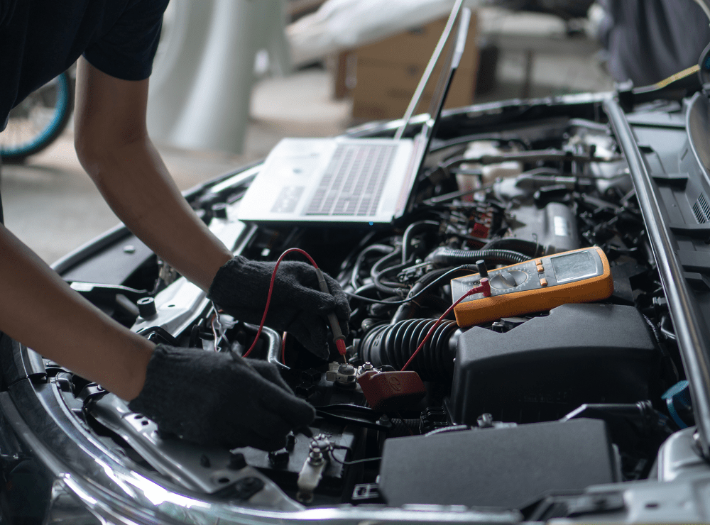 A mechanic wearing gloves is diagnosing a car engine using a multimeter.